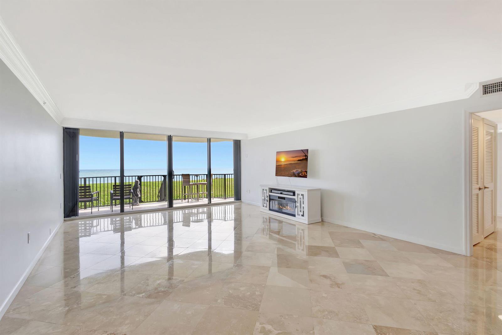 A bright living room features polished tile floors, a wall-mounted television, and a balcony view — 400 Beach Road # 103, Tequesta FL 33469, TEQUESTA, FL