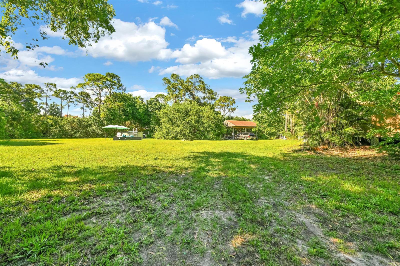 A large, grassy backyard features mature trees, a covered carport, and a patio umbrella — 17605 92nd Lane N, FL 33470, FL