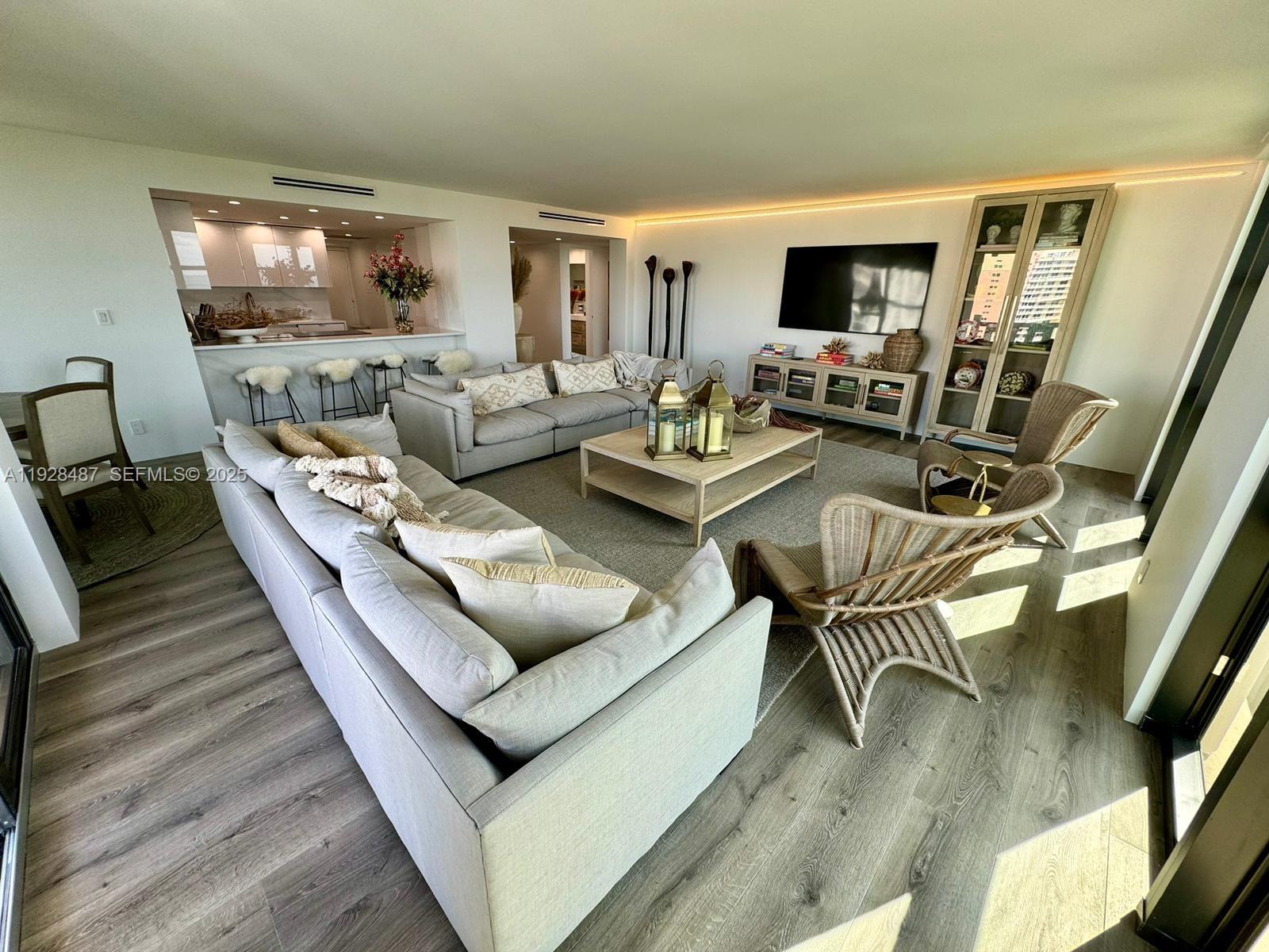 A spacious living room features light wood flooring, neutral sofas, wicker chairs, and a kitchen — 1121 Crandon Blvd # E1001, Key Biscayne FL 33149, KEY BISCAYNE, FL