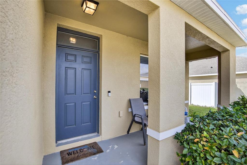 A covered front porch features a blue door, a welcome mat, and a single chair — 35716 HILLBROOK AVENUE, ZEPHYRHILLS, FL