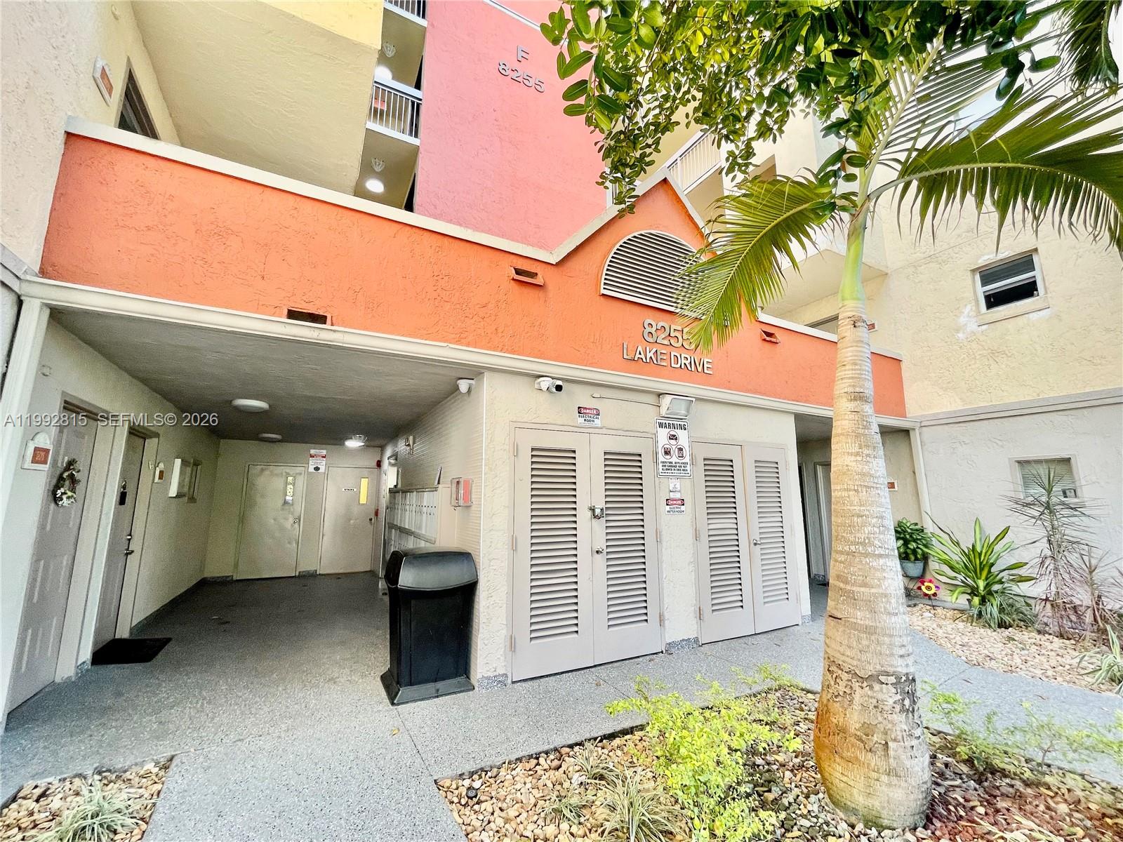 An exterior view of a multi-story apartment building entrance with a palm tree and walkway — 8255 Lake Dr # 102, Doral FL 33166, DORAL, FL