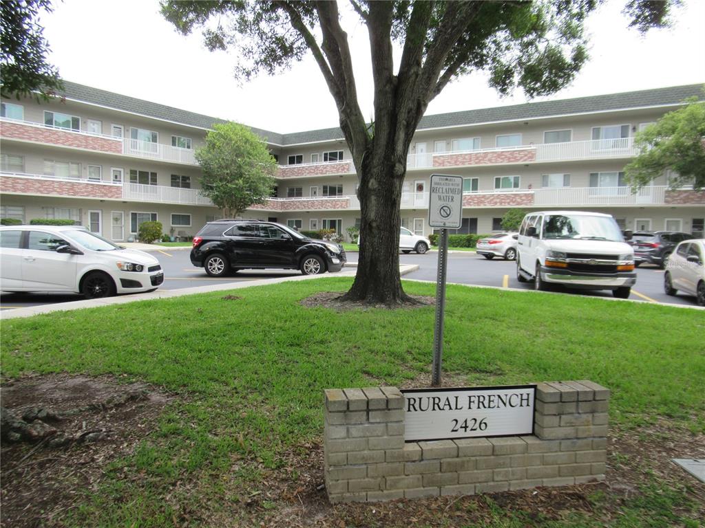 A three-story apartment building with a parking lot and a sign in the foreground — 2426 PERSIAN DRIVE UNIT 56, CLEARWATER, FL