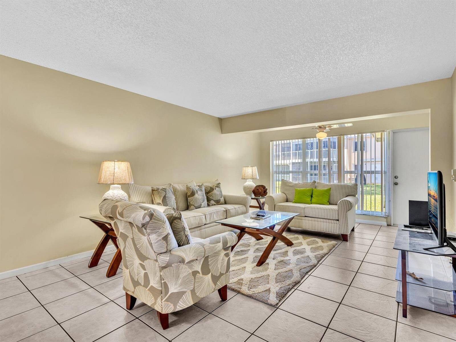 A living room features tile flooring, a patterned armchair, two sofas, and a glass-topped table — 1610 NW 18th Avenue # 96d, Delray Beach FL 33445, DELRAY BEACH, FL