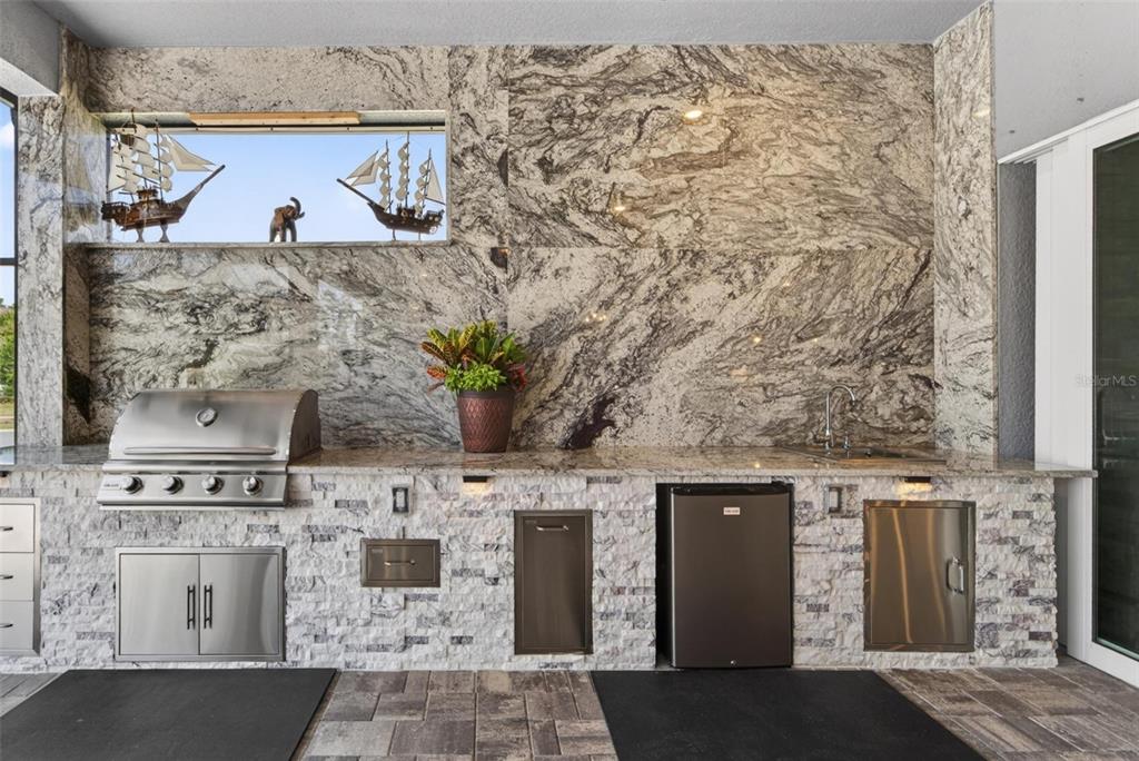 An outdoor kitchen features a stone backsplash, granite countertop, stainless steel grill, and small refrigerator — 18752 HORIZON VIEW POINT, LAND O LAKES, FL