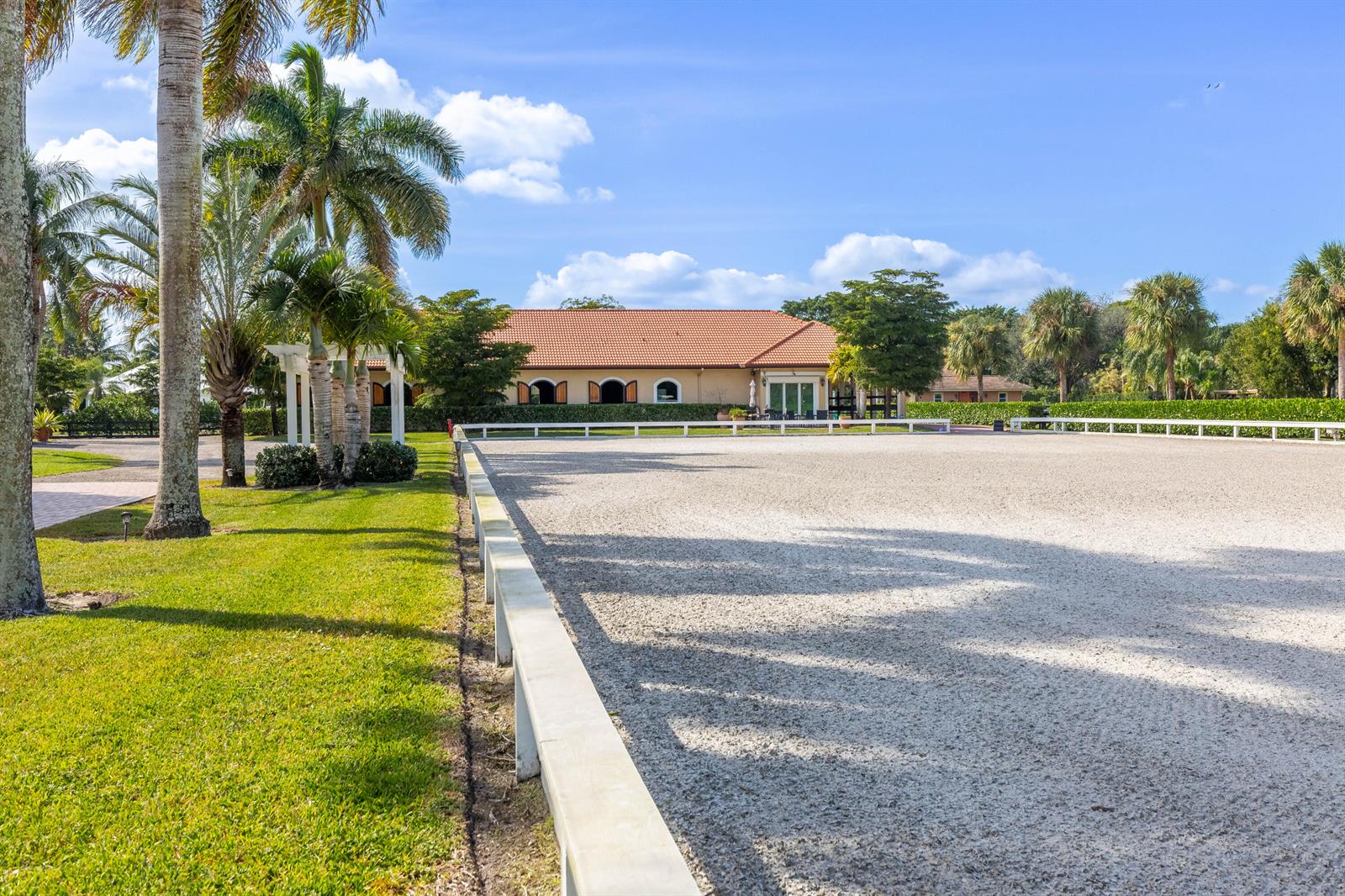 A large, sandy horse riding arena sits in front of a house with palm trees — 14943 Paddock Drive, Wellington FL 33414, WELLINGTON, FL