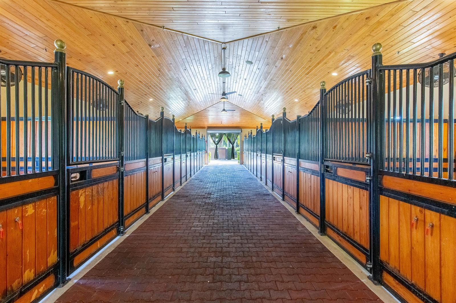 A horse stable aisle features brick flooring, wood-paneled ceilings, and black metal stall doors — 14943 Paddock Drive, Wellington FL 33414, WELLINGTON, FL