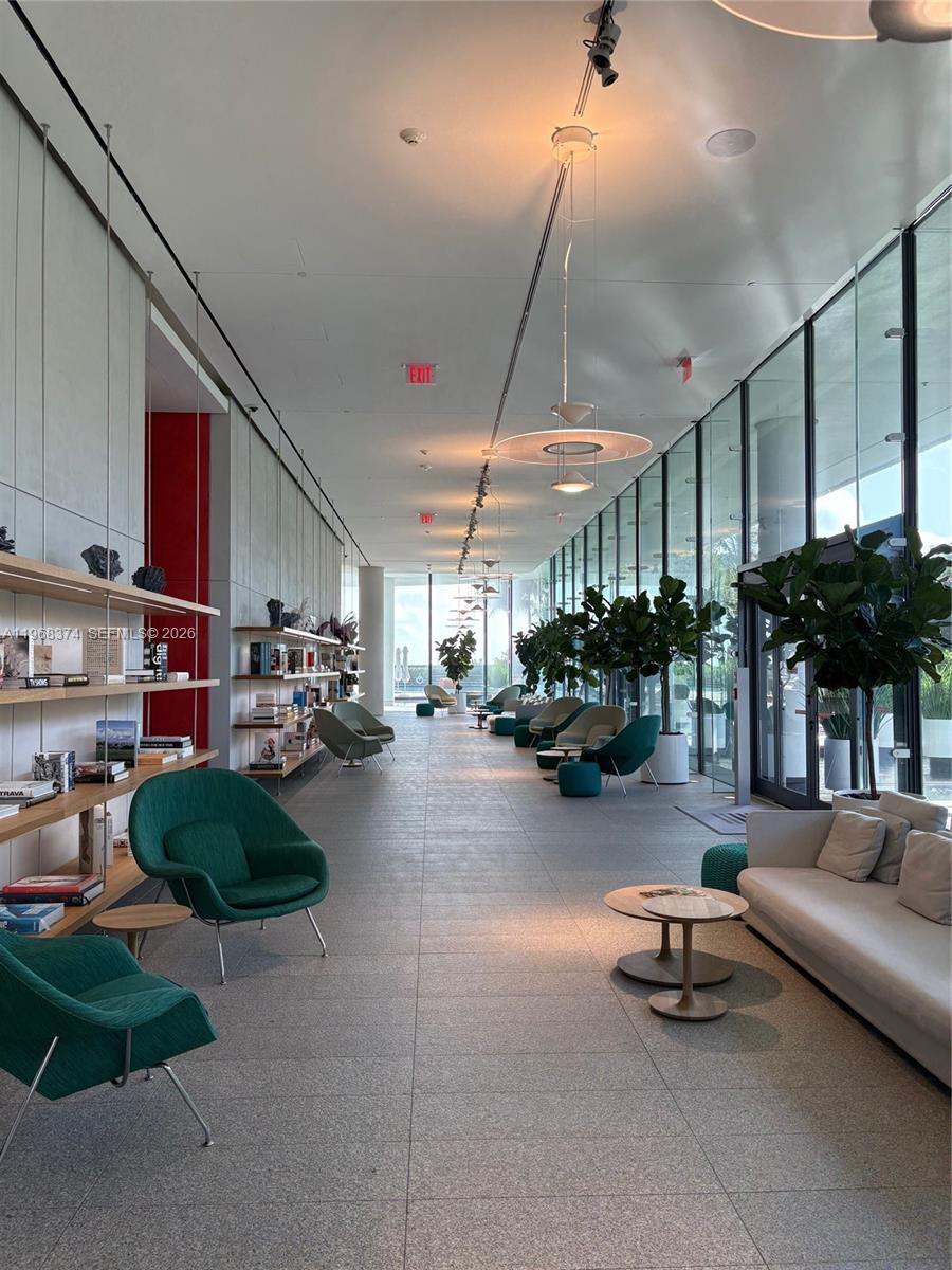 A long, modern lounge area features green armchairs, bookshelves, large potted plants, and floor-to-ceiling windows — 8701 Collins Ave # 803, Miami Beach FL 33154, MIAMI BEACH, FL