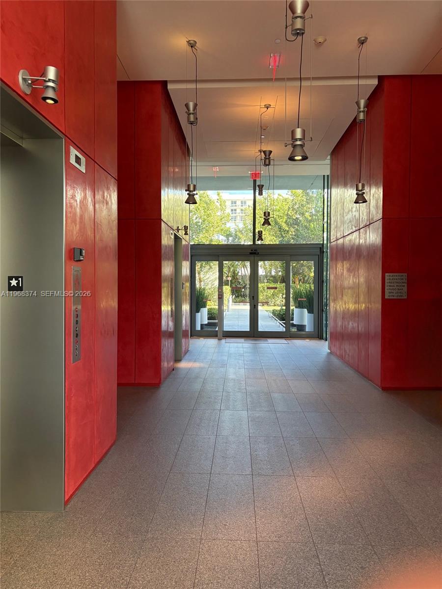 A modern building lobby features red paneled walls, tile flooring, and glass doors leading outside — 8701 Collins Ave # 803, Miami Beach FL 33154, MIAMI BEACH, FL