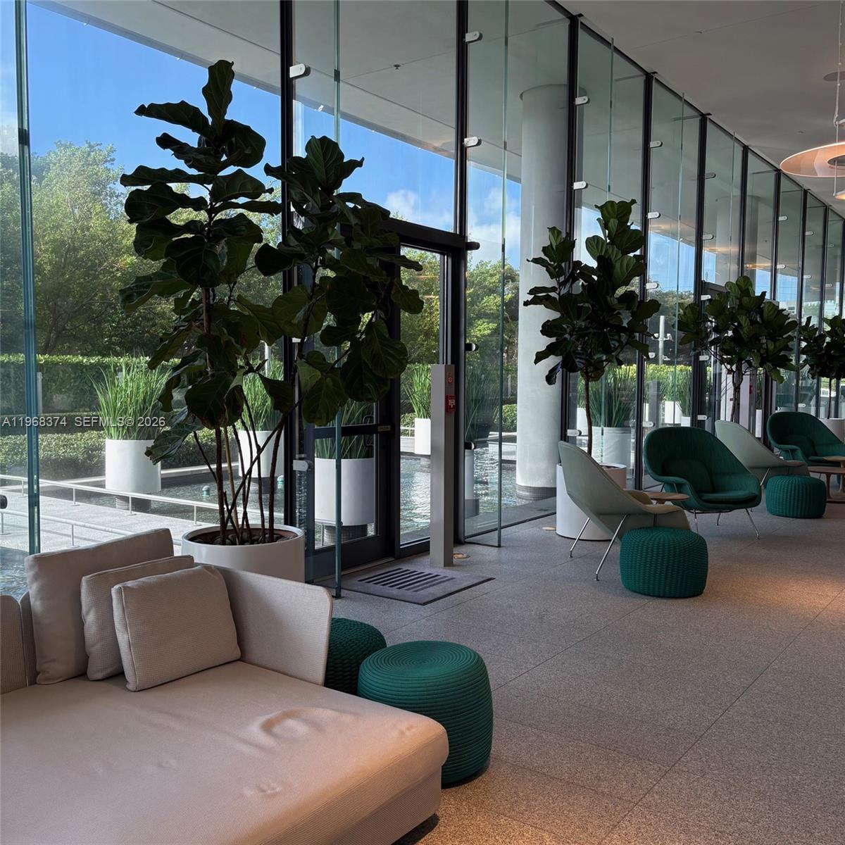 A modern lobby features floor-to-ceiling glass walls, potted fiddle-leaf fig trees, and contemporary lounge seating — 8701 Collins Ave # 803, Miami Beach FL 33154, MIAMI BEACH, FL