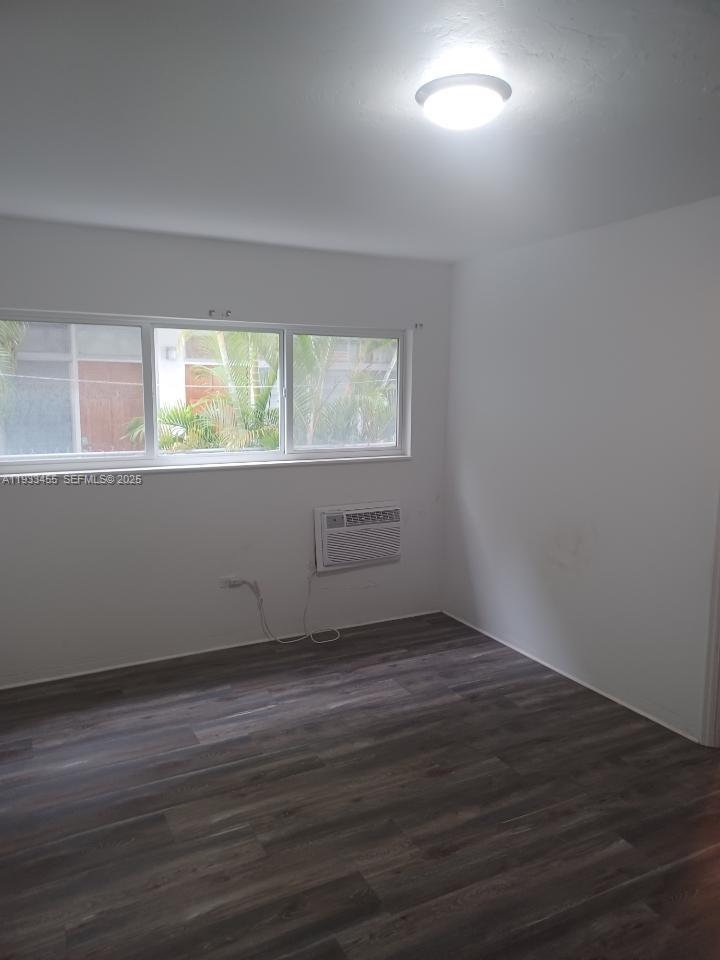 An empty room features white walls, dark wood-look flooring, a window, and a wall-mounted air conditioner — 6950 Byron Ave # 3, Miami Beach FL 33141, MIAMI BEACH, FL