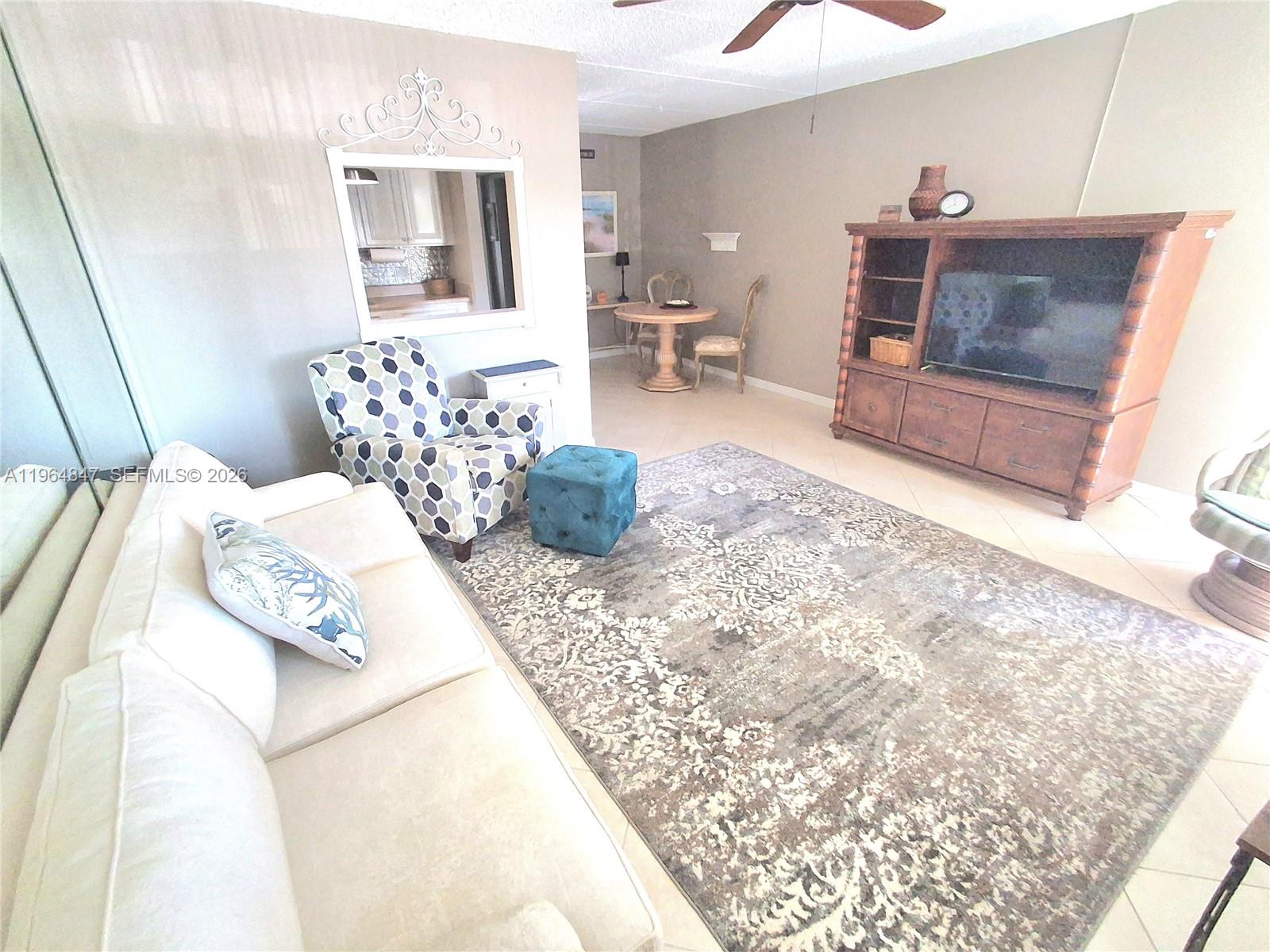 A living room features a white sofa, patterned armchair, large area rug, and wooden entertainment center — 3000 Springdale Blvd # V207, Palm Springs FL 33461, PALM SPRINGS, FL