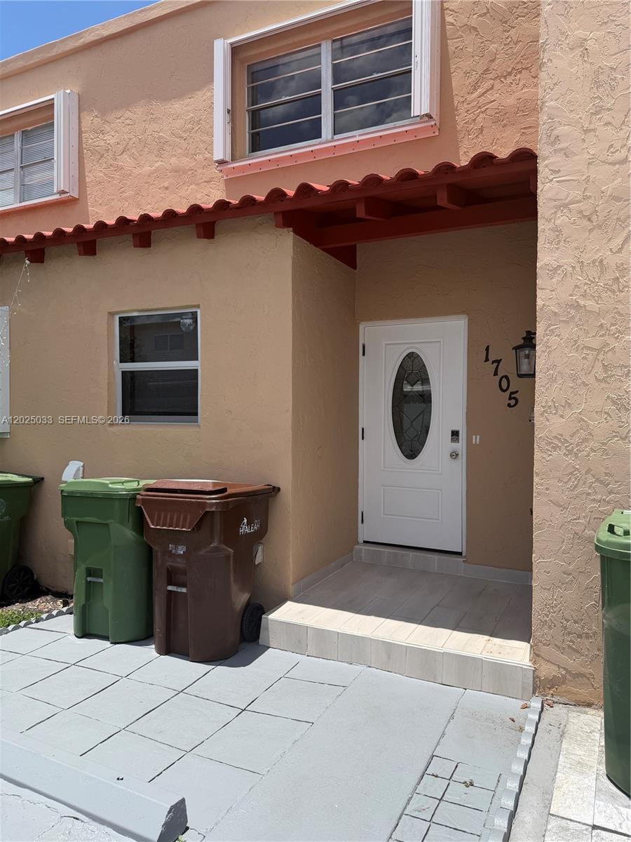 The exterior of a tan stucco building features a white front door and trash bins — 1705 W 57th St # 506, Hialeah FL 33012, HIALEAH, FL