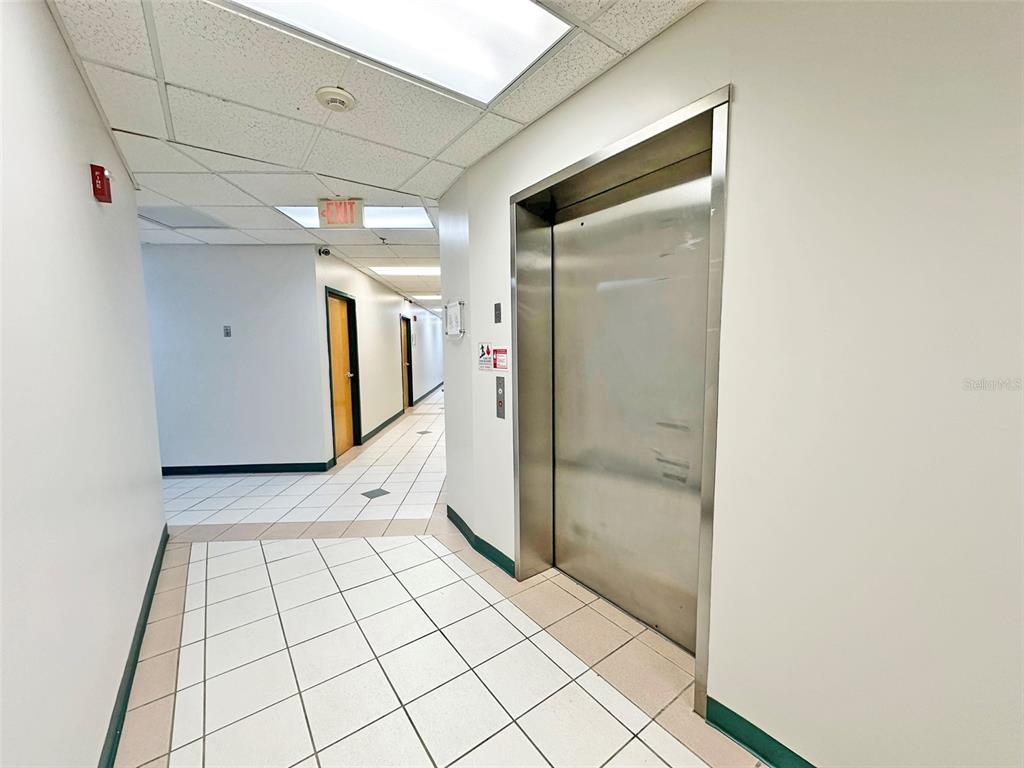 A hallway with white tiled floors leads past an elevator to a corridor with doors — 7802 KINGSPOINTE PARKWAY UNIT 210, ORLANDO, FL