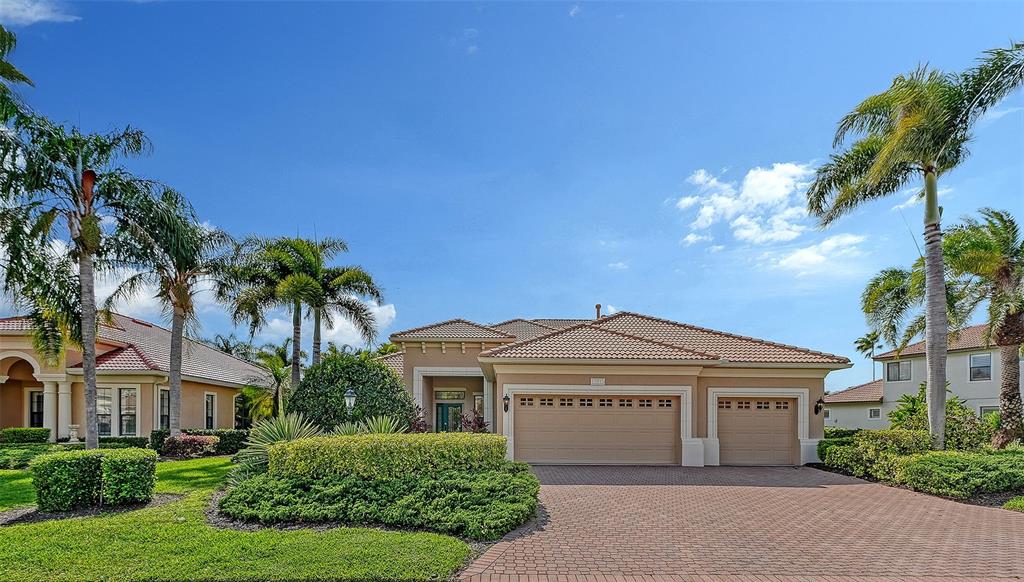 A tan single-story house with a three-car garage and palm trees under a blue sky — 13911 SIENA LOOP, LAKEWOOD RANCH, FL