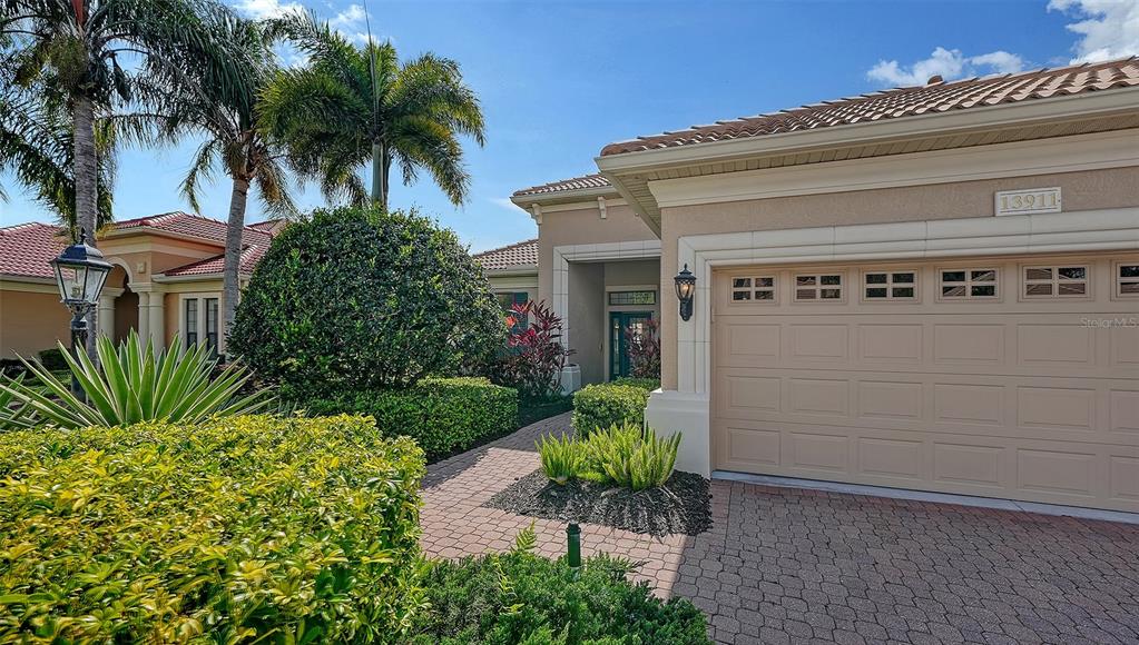 A beige house exterior features a two-car garage, a paver driveway, and landscaped front gardens — 13911 SIENA LOOP, LAKEWOOD RANCH, FL