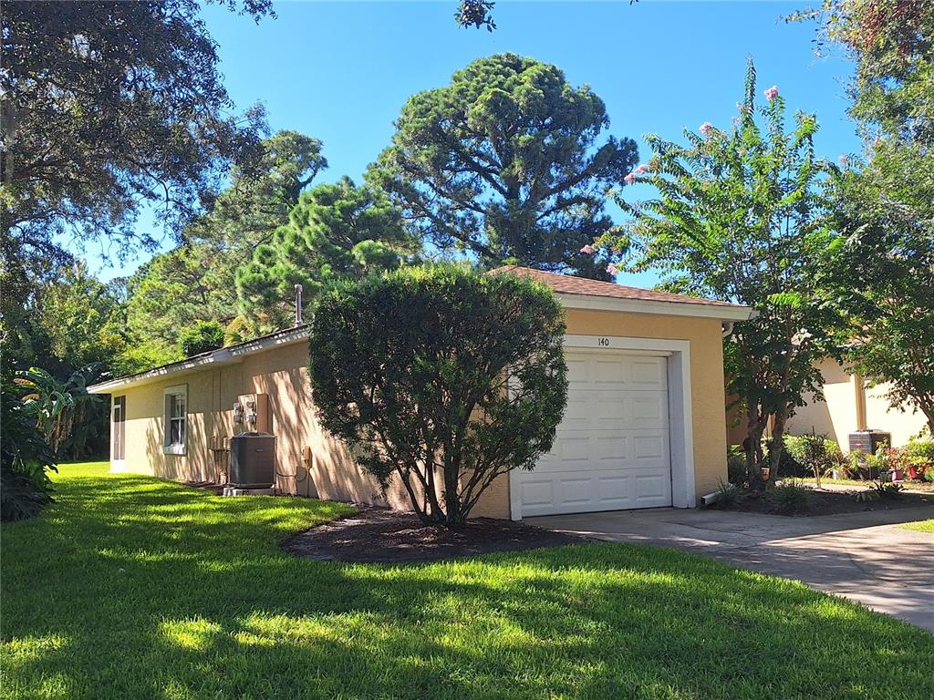 A single-story house with a white garage door, tan stucco walls, and surrounding trees — 140 TURNBULL VILLAS CIRCLE, NEW SMYRNA BEACH, FL