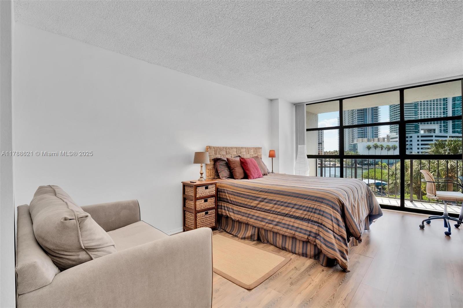 A bedroom features a bed, a beige armchair, and floor-to-ceiling windows overlooking a city skyline — 600 Parkview Dr, Hallandale Beach FL 33009, HALLANDALE BEACH, FL