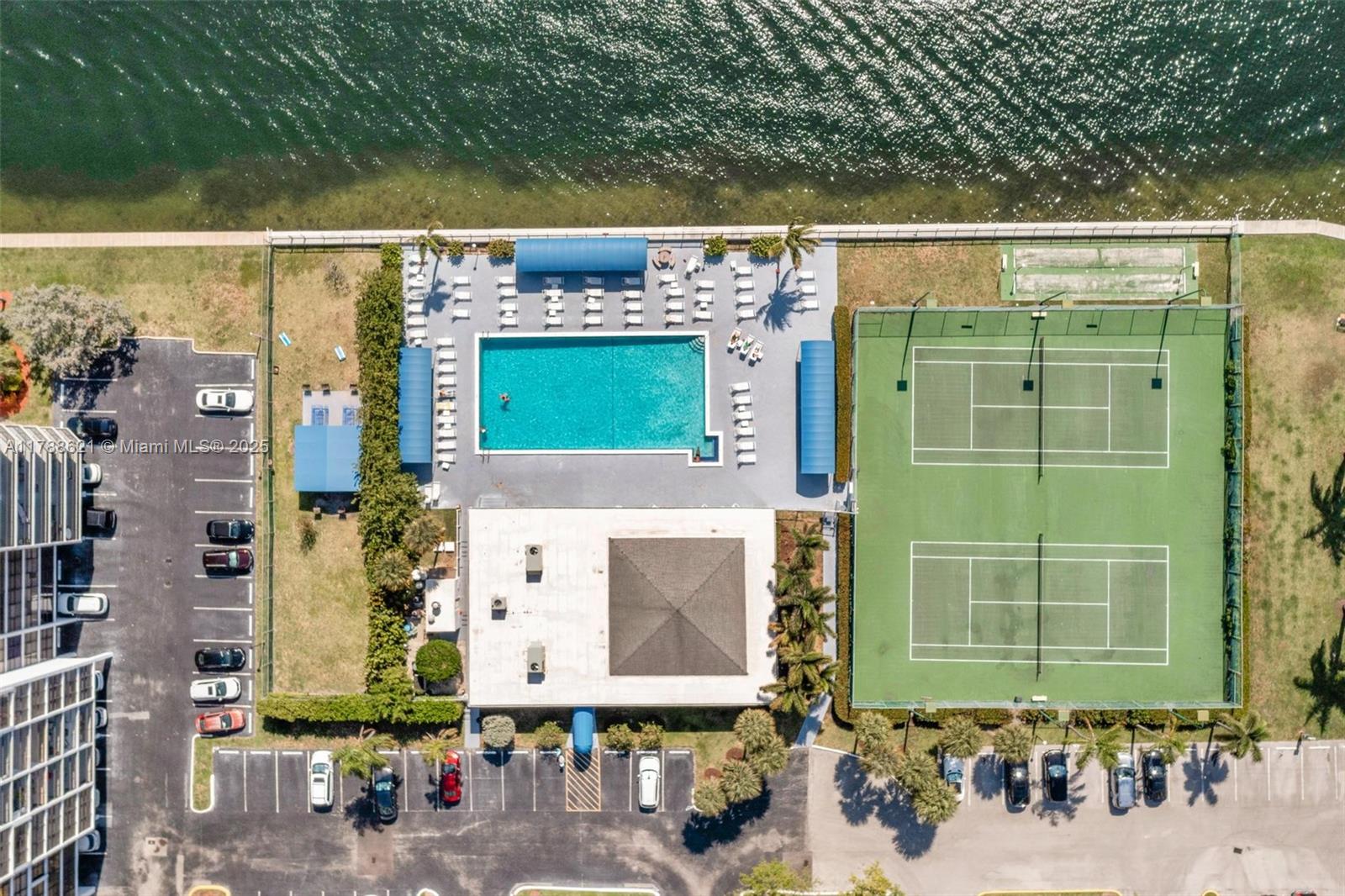An aerial view shows a swimming pool, two tennis courts, and parking lots near water — 600 Parkview Dr, Hallandale Beach FL 33009, HALLANDALE BEACH, FL