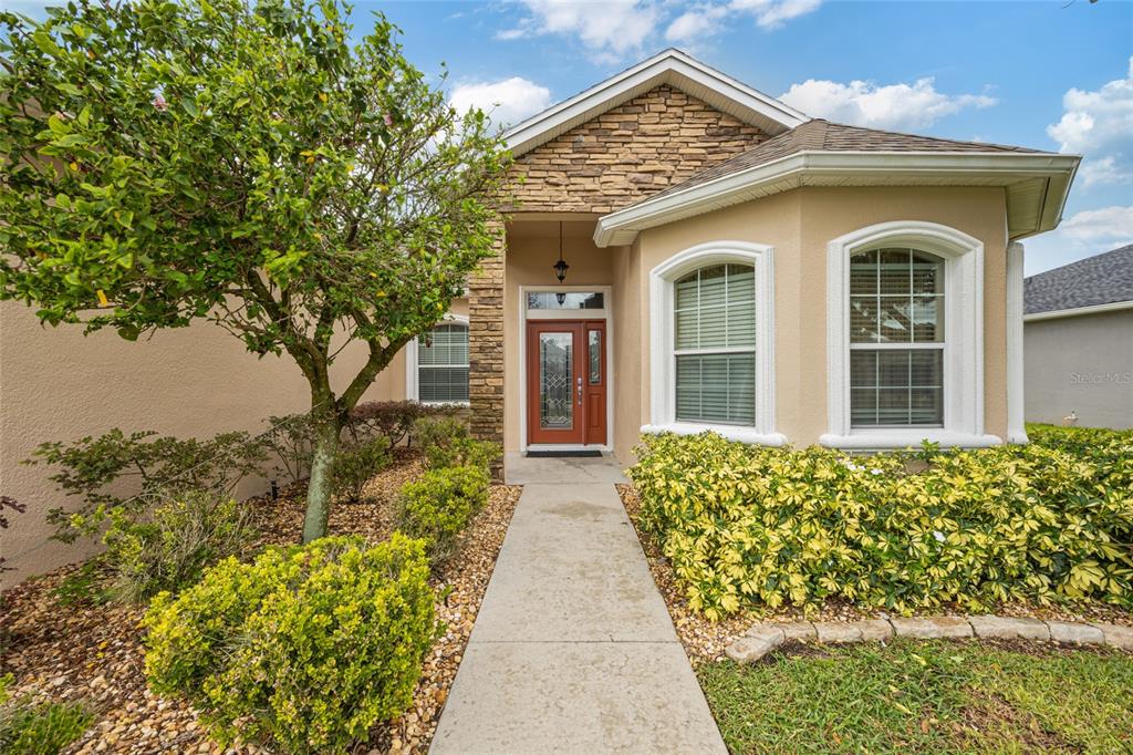 A tan house exterior features a stone-accented entryway, a red front door, and landscaped gardens — 2836 WHITNEY STREET, LAKELAND, FL
