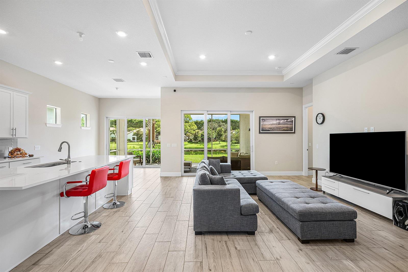 An open-concept living room and kitchen with gray furniture, red bar stools, and sliding doors — 349 Geillis Path, Delray Beach FL 33445, DELRAY BEACH, FL