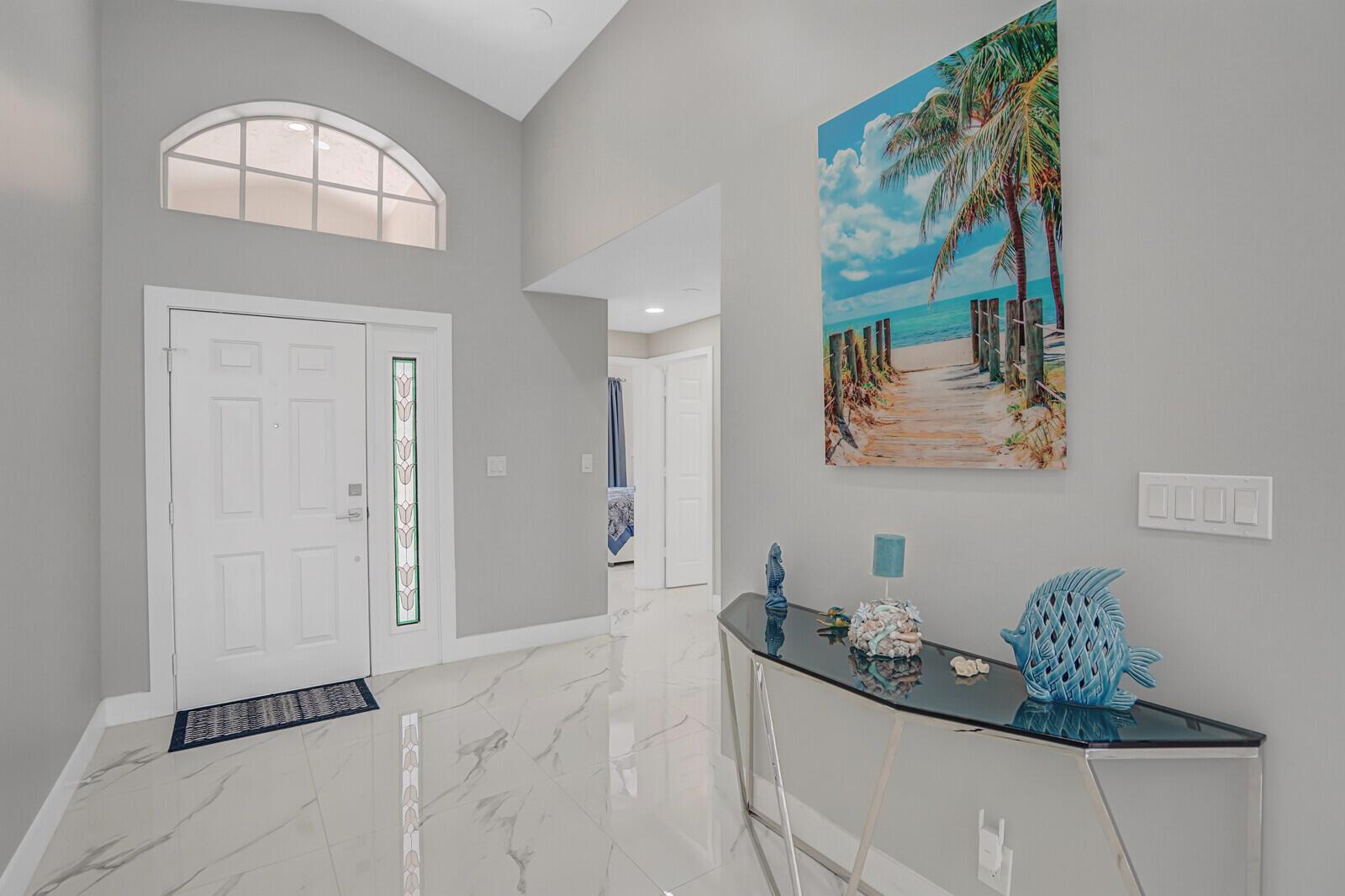 A bright entryway features white marble floors, a glass console table, and a beach-themed painting — 7791 Great Glen Circle, Delray Beach FL 33446, DELRAY BEACH, FL