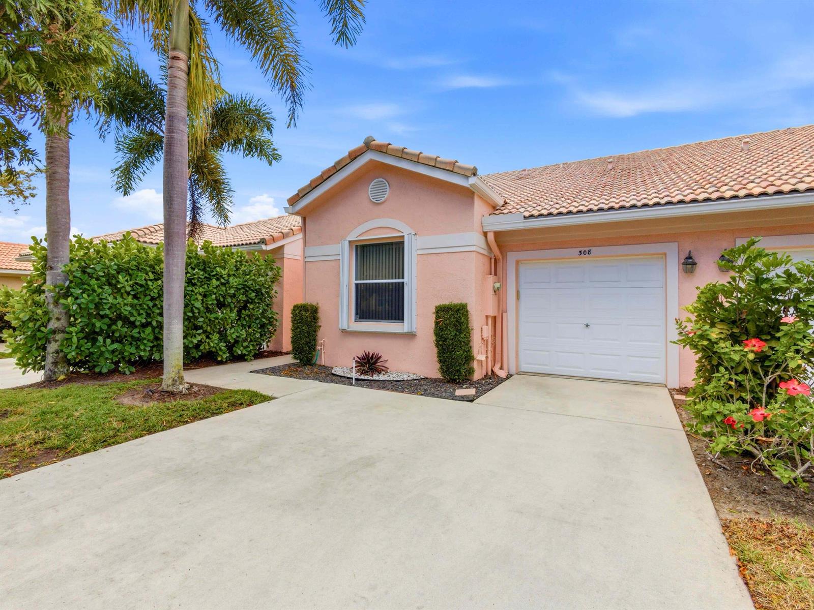 A pink single-story house features a white garage door, a concrete driveway, and landscaping — 308 Coral Trace Court, Delray Beach FL 33445, DELRAY BEACH, FL