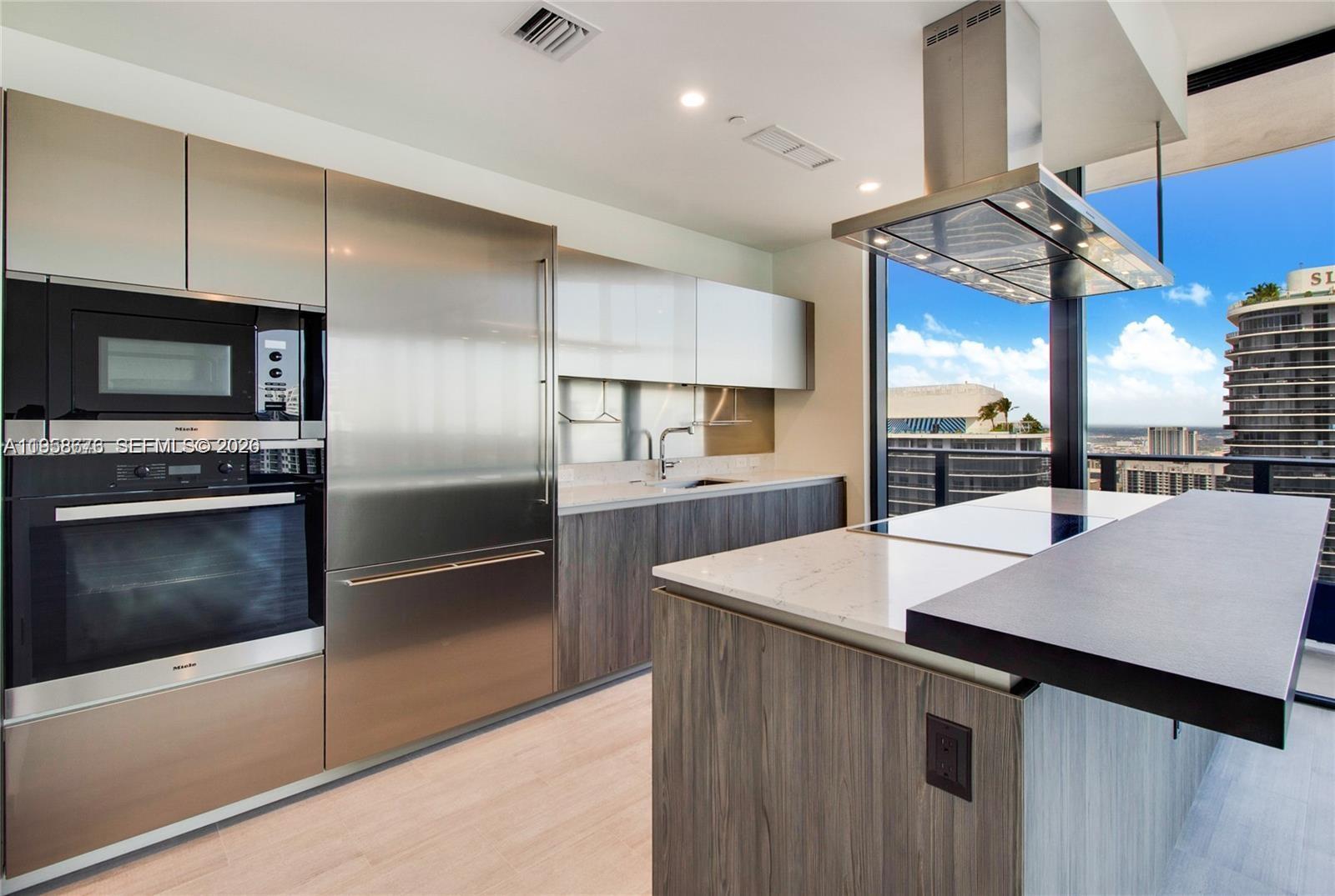 A modern kitchen features stainless steel appliances, wood-grain cabinetry, and a large island with views — 1000 Brickell Plz, Miami FL 33131, MIAMI, FL