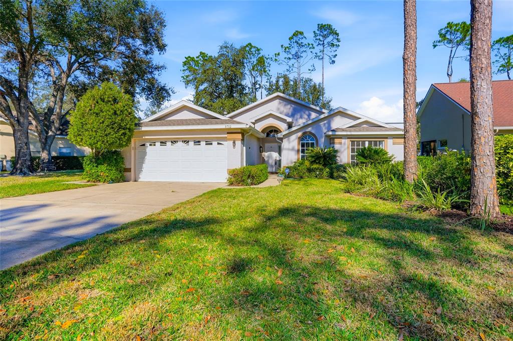 A single-story house with a white garage, beige exterior, and a grassy front lawn — 5712 WHITEBARK DRIVE, WESLEY CHAPEL, FL