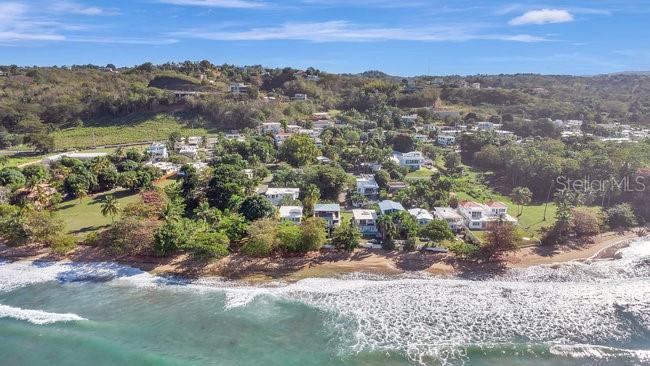 An aerial view shows houses nestled along a coastline with trees and ocean waves — 40 CALLE SURF BO ENSENADA, RINCON, PR