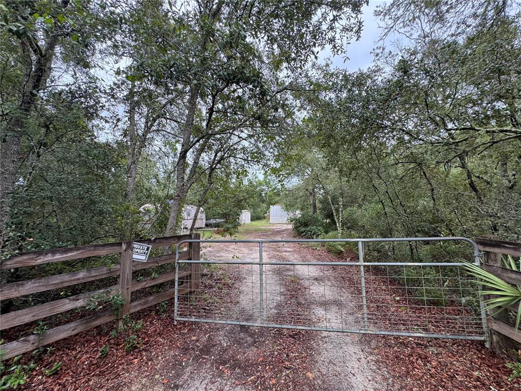 A metal gate blocks a dirt driveway leading to a wooded lot with small structures — 23180 NE 156TH COURT, FORT MC COY, FL