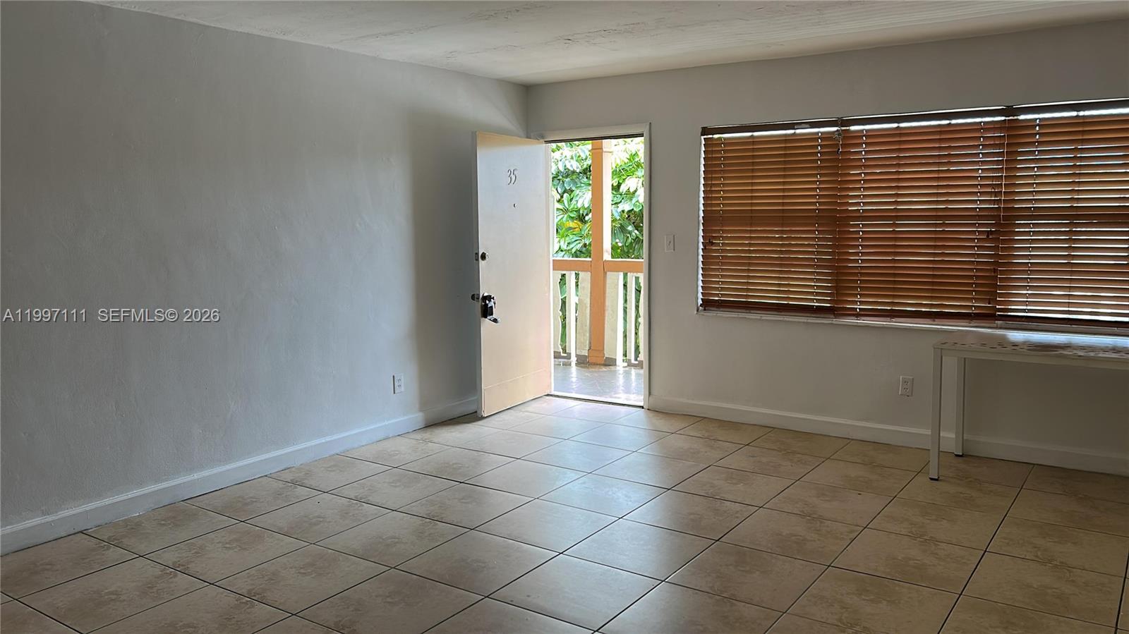 An empty room features beige tile flooring, white walls, a doorway to a balcony, and blinds — 1855 NE 121st St # 35, North Miami FL 33181, NORTH MIAMI, FL
