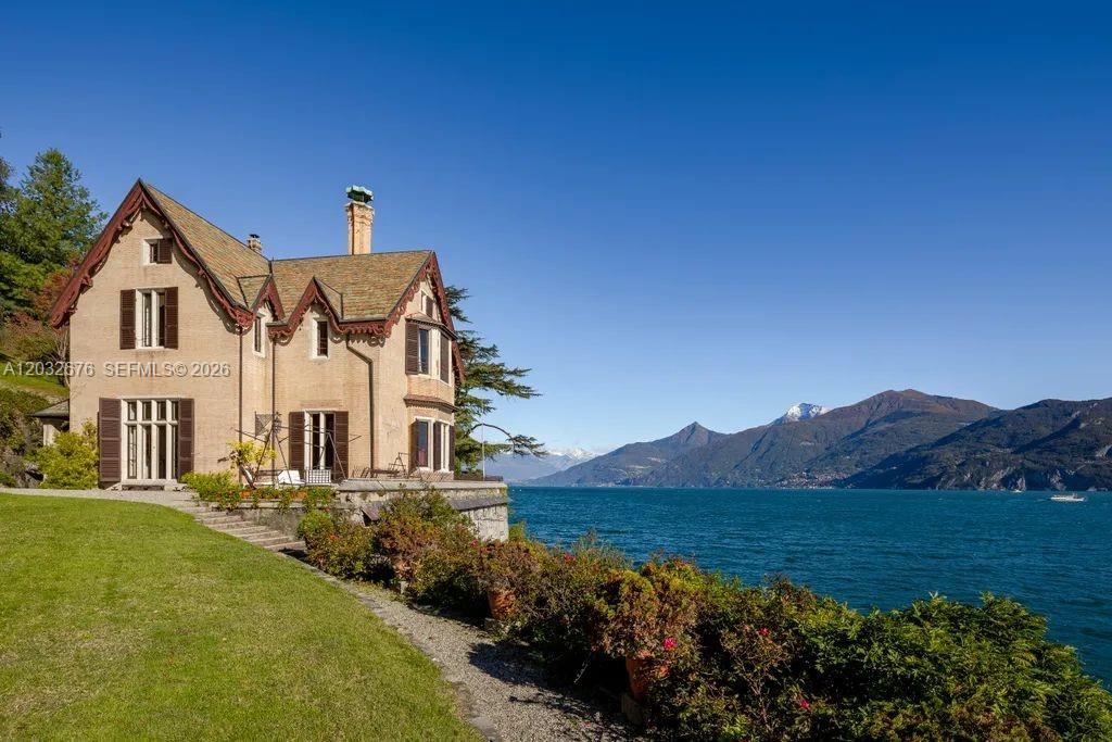 A multi-story house with brown shutters sits on a grassy lawn beside a lake — Lake Como Luxury Real Estate for Sale Max Mlekus, FL