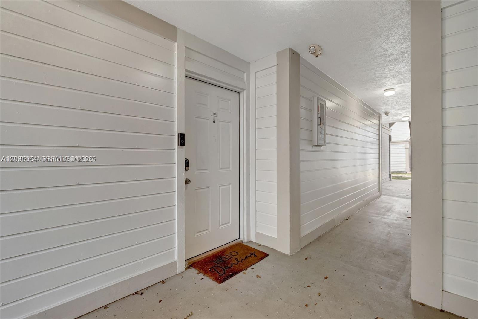 A white exterior door with a welcome mat is situated in a covered outdoor hallway — 18965 NW 62nd Ave # 210, Hialeah FL 33015, HIALEAH, FL
