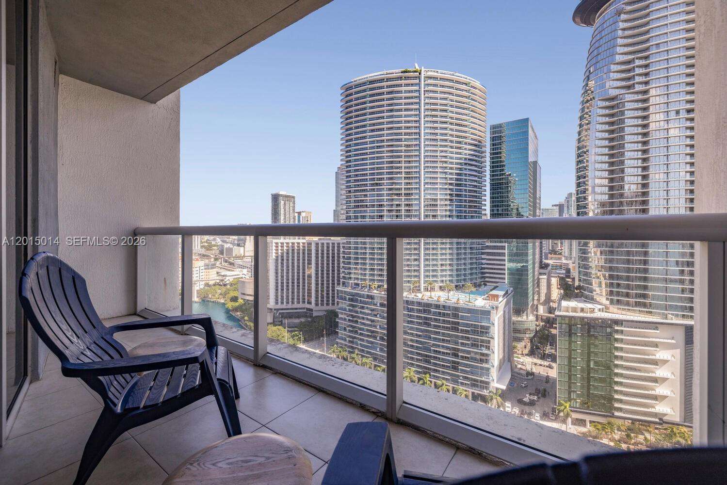 A balcony with a dark chair overlooks a dense urban skyline of tall glass skyscrapers — 475 Brickell Ave # 2610, Miami FL 33131, MIAMI, FL