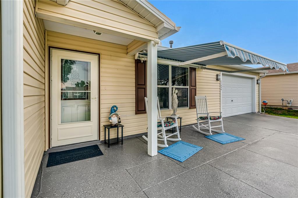 A front porch with two rocking chairs, a blue awning, and a cream-colored house exterior — 17654 SE 93RD BUTLER COURT, THE VILLAGES, FL