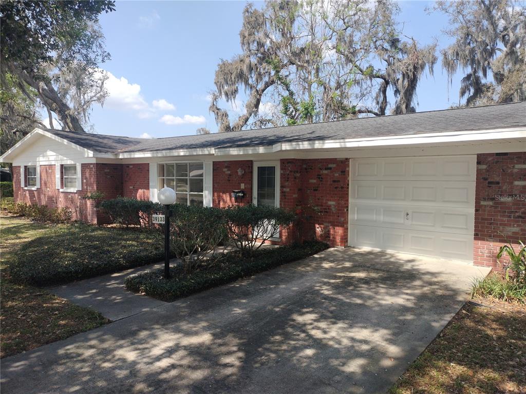 A single-story brick house features a white garage door and a front yard with trees — 39133 5TH AVENUE, ZEPHYRHILLS, FL