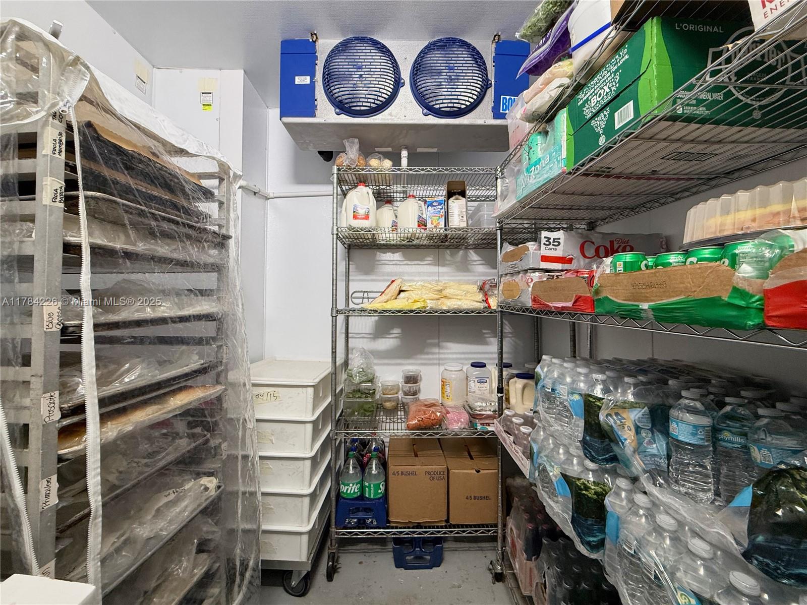 A walk-in refrigerator contains metal shelving units stocked with food, beverages, and plastic storage bins — E Las Olas Blvd, Fort Lauderdale FL 33316, FORT LAUDERDALE, FL