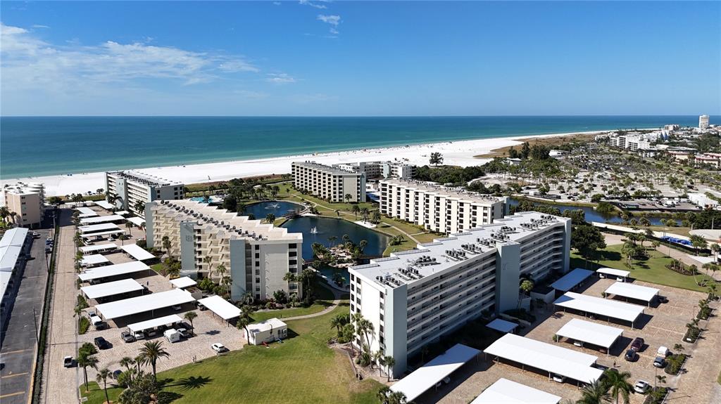 An aerial view shows several multi-story apartment buildings surrounding a central pond near a beach — 5770 MIDNIGHT PASS ROAD UNIT 301, SARASOTA, FL
