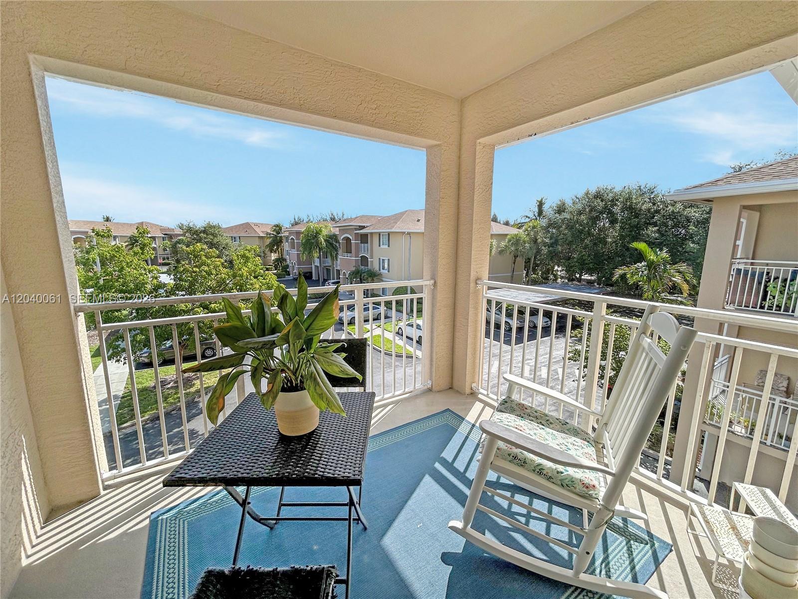 A balcony features a blue rug, a small table with a plant, and a rocking chair — 6458 Emerald Dunes Dr # 303, West Palm Beach FL 33411, WEST PALM BEACH, FL