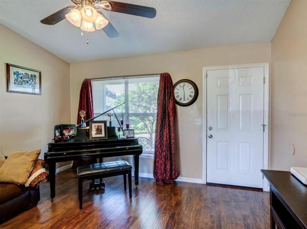 A room with wood-look flooring features a black piano, a window with curtains, and a door — 1730 TROPICAL COURT, TAVARES, FL