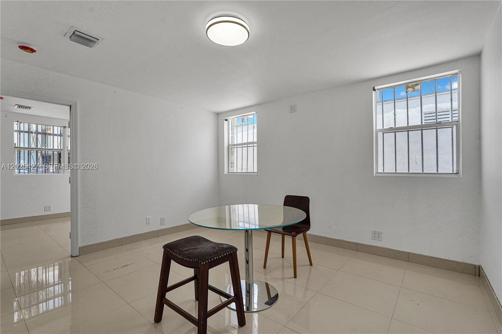 A sparsely furnished room with white walls, tile floors, two windows with security bars, and a table — 7525 NE 3rd Pl, Miami FL 33138, MIAMI, FL