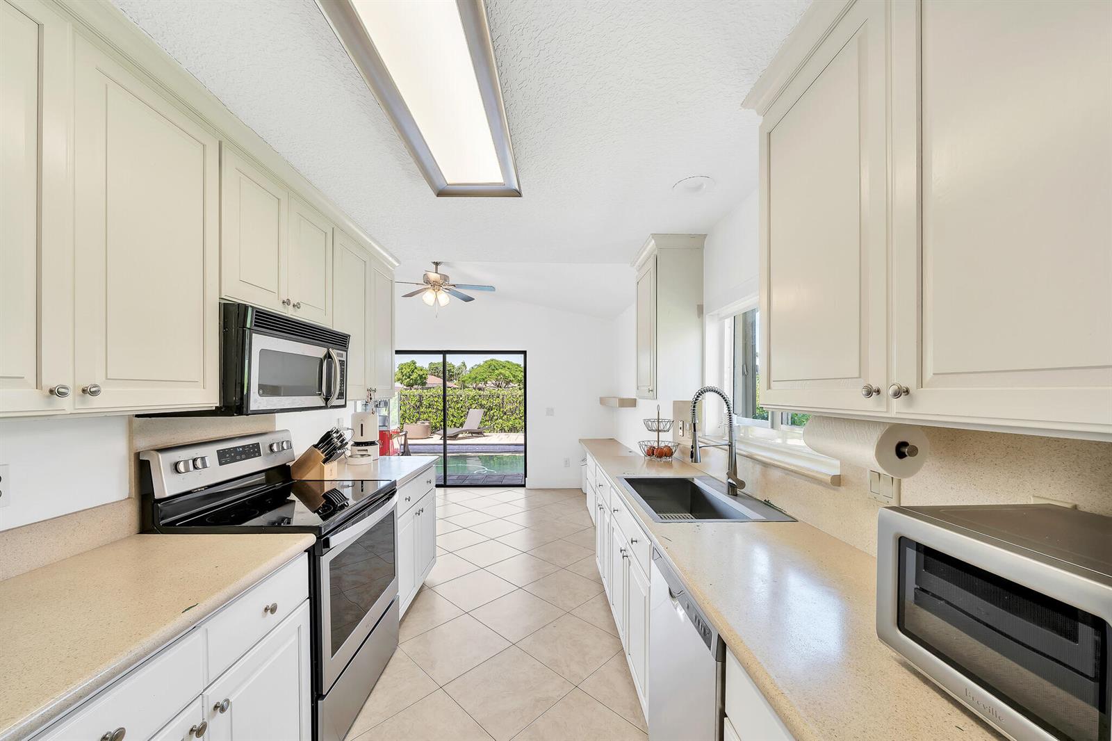 A galley kitchen features white cabinets, beige countertops, stainless steel appliances, and a backyard pool view — 10255 Sunset Bend Drive, Boca Raton FL 33428, BOCA RATON, FL
