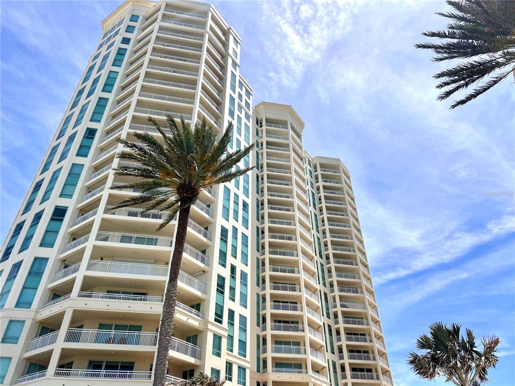 A low-angle view of a tall, multi-story residential building with balconies and palm trees — 1180 GULF BLVD UNIT 306, CLEARWATER, FL