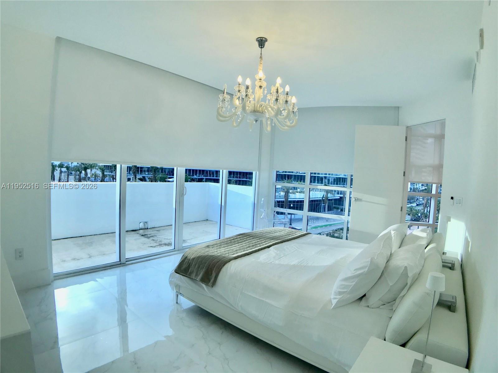 A modern bedroom features a white bed, marble floors, a crystal chandelier, and floor-to-ceiling windows — 50 S Pointe Dr, Miami Beach FL 33139, MIAMI BEACH, FL