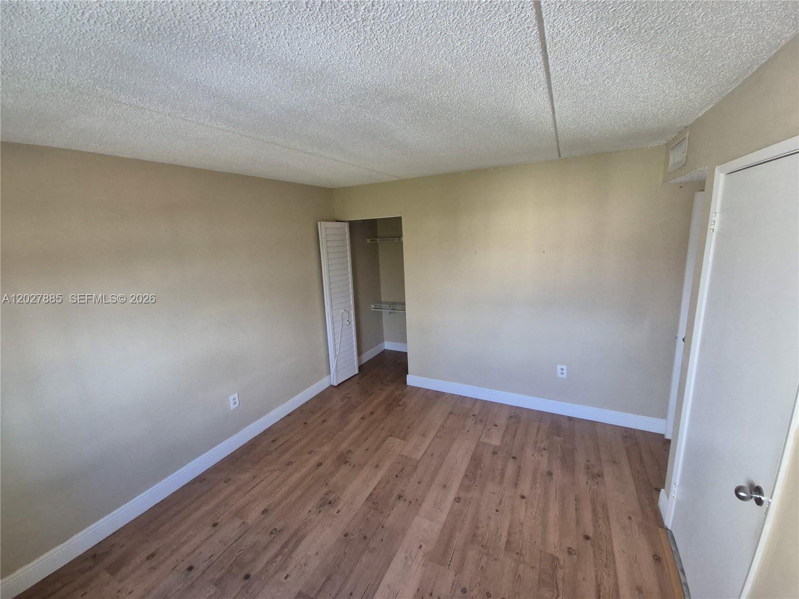 An empty bedroom features beige walls, wood-look flooring, a popcorn ceiling, and a small closet — 6045 NW 186th St # 308, Hialeah FL 33015, HIALEAH, FL