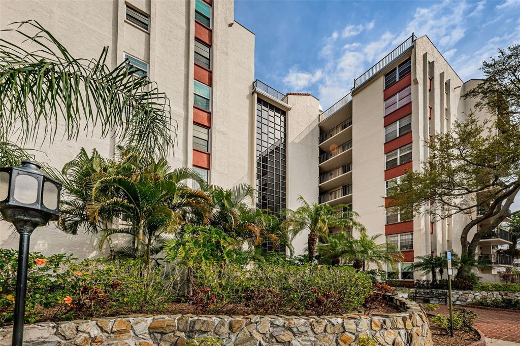 A multi-story apartment building features a stone retaining wall and palm trees in front — 2699 SEVILLE BOULEVARD UNIT 105, CLEARWATER, FL