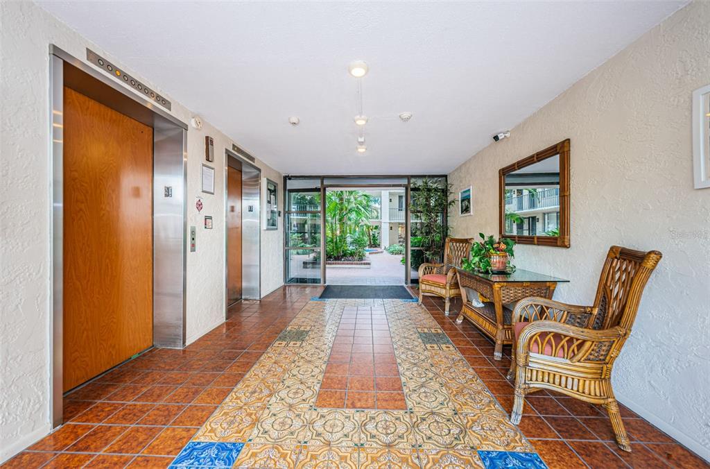 An indoor building lobby features tiled floors, two elevators, and wicker chairs near a glass table — 2699 SEVILLE BOULEVARD UNIT 105, CLEARWATER, FL