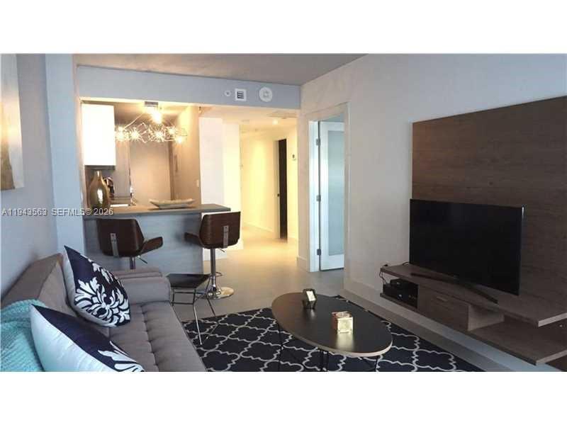A living room with a grey sofa, a TV on a wood panel, and a kitchen — 100 Lincoln Rd # 420, Miami Beach FL 33139, MIAMI BEACH, FL