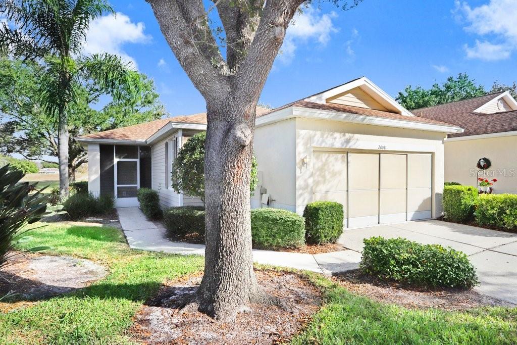 A single-story house with a beige exterior, a two-car garage, and a large front tree — 2608 NEWCOMB COURT UNIT 2608, SUN CITY CENTER, FL