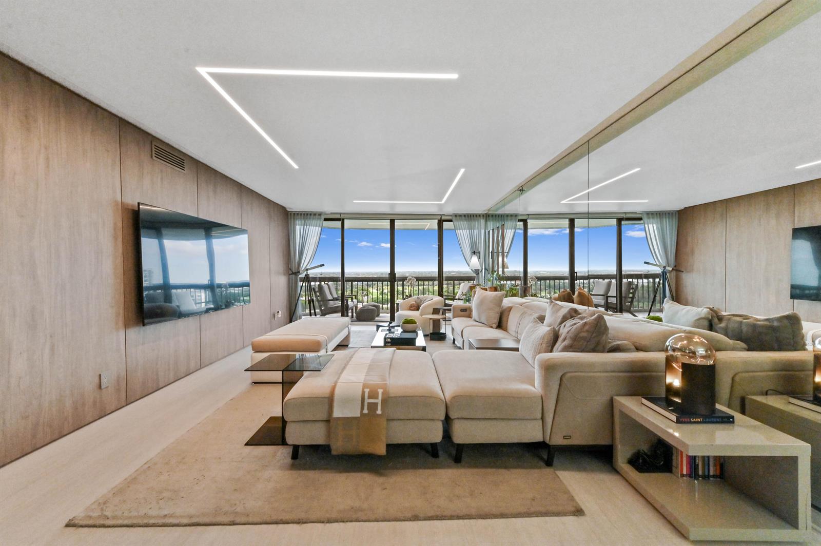 A modern living room features wood-paneled walls, a large sectional sofa, and floor-to-ceiling windows — 1900 Consulate Place # 1205, West Palm Beach FL 33401, WEST PALM BEACH, FL